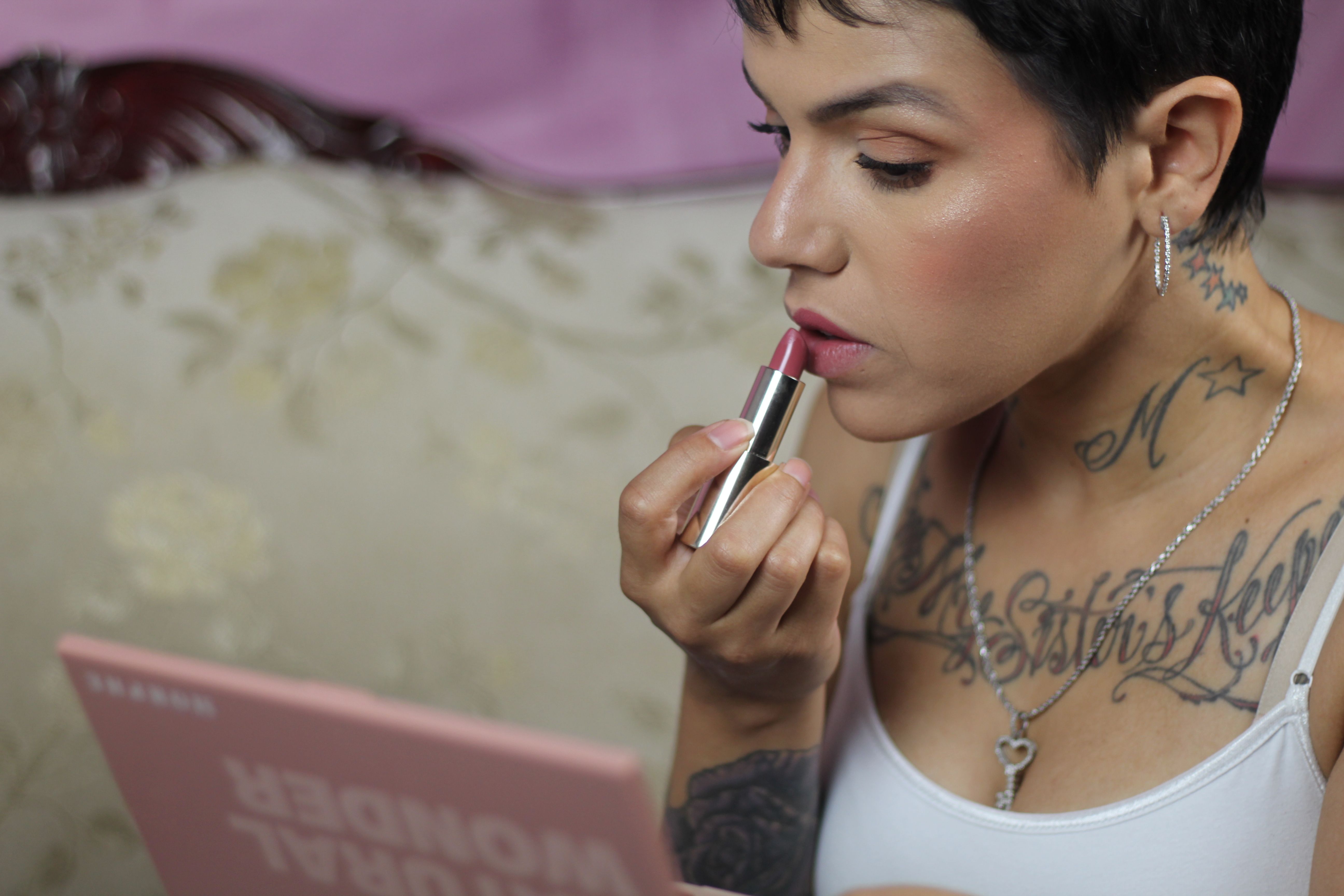 Close-up of a woman applying red lipstick, showcasing tattoos and makeup artistry in a cozy indoor setting.