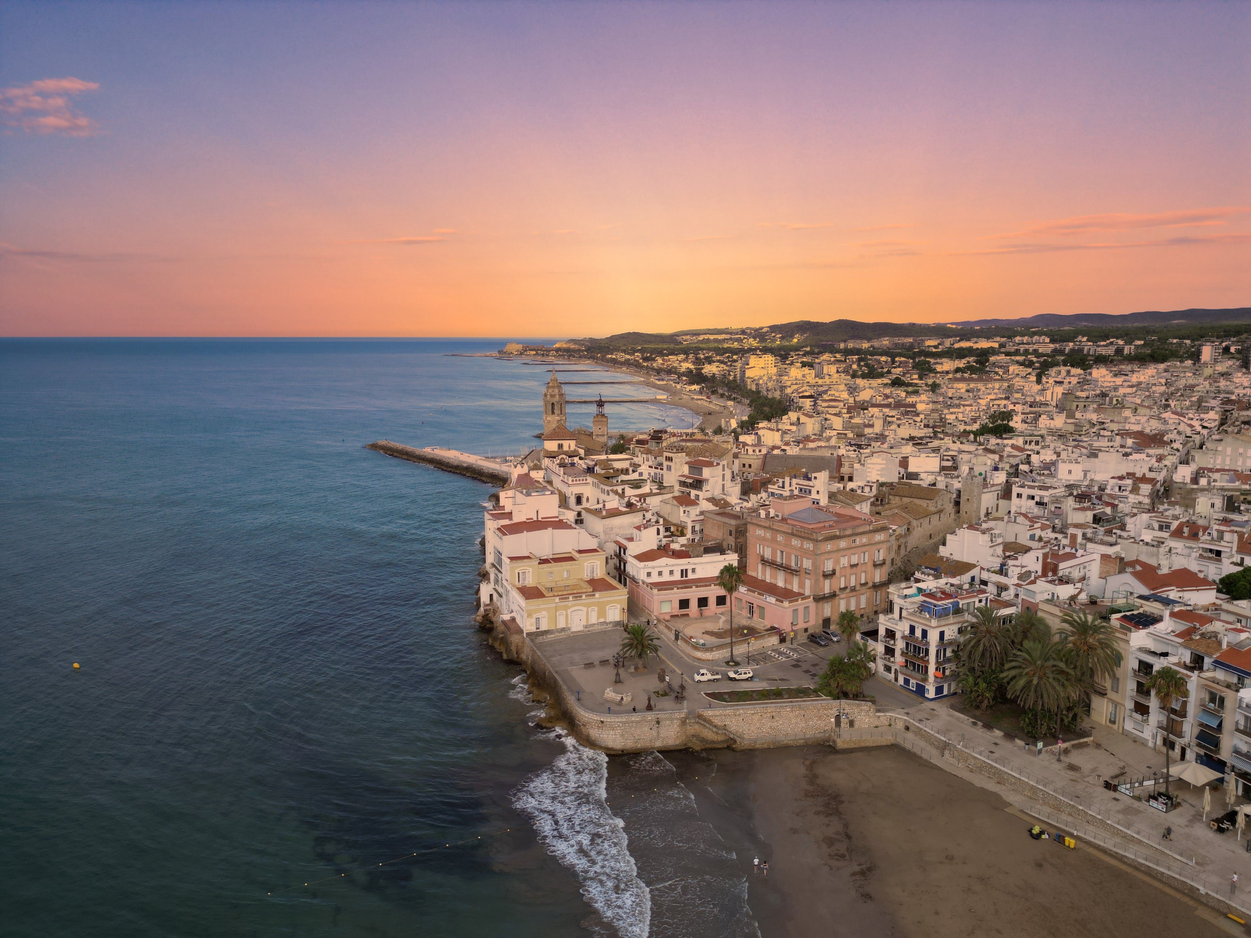 A coastal town curves around the sea beneath a pink and orange sunset sky