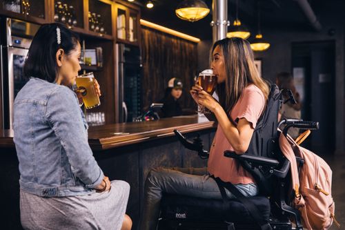 Free Two women enjoying beer at a bar, one seated in a wheelchair, capturing a moment of camaraderie. Stock Photo