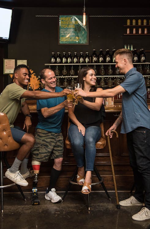 Foto de quatro amigos bebendo no balcão de um bar. Estão brindando e rindo, temos uma pessoa preta, uma pessoa com deficiencia usando protese na perna, uma mulher de pele branca e um homem de pele branca com uma bengala. Todos seguram um copo de cerveja cheio