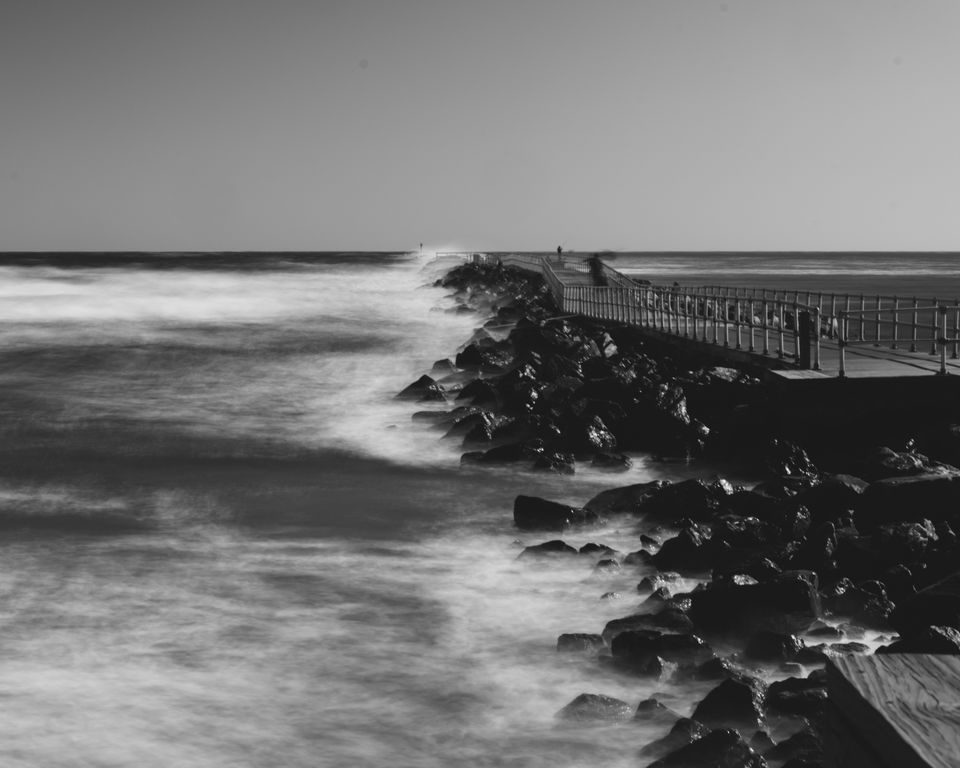 Drammatica foto in bianco e nero di onde oceaniche che si infrangono contro un molo roccioso.