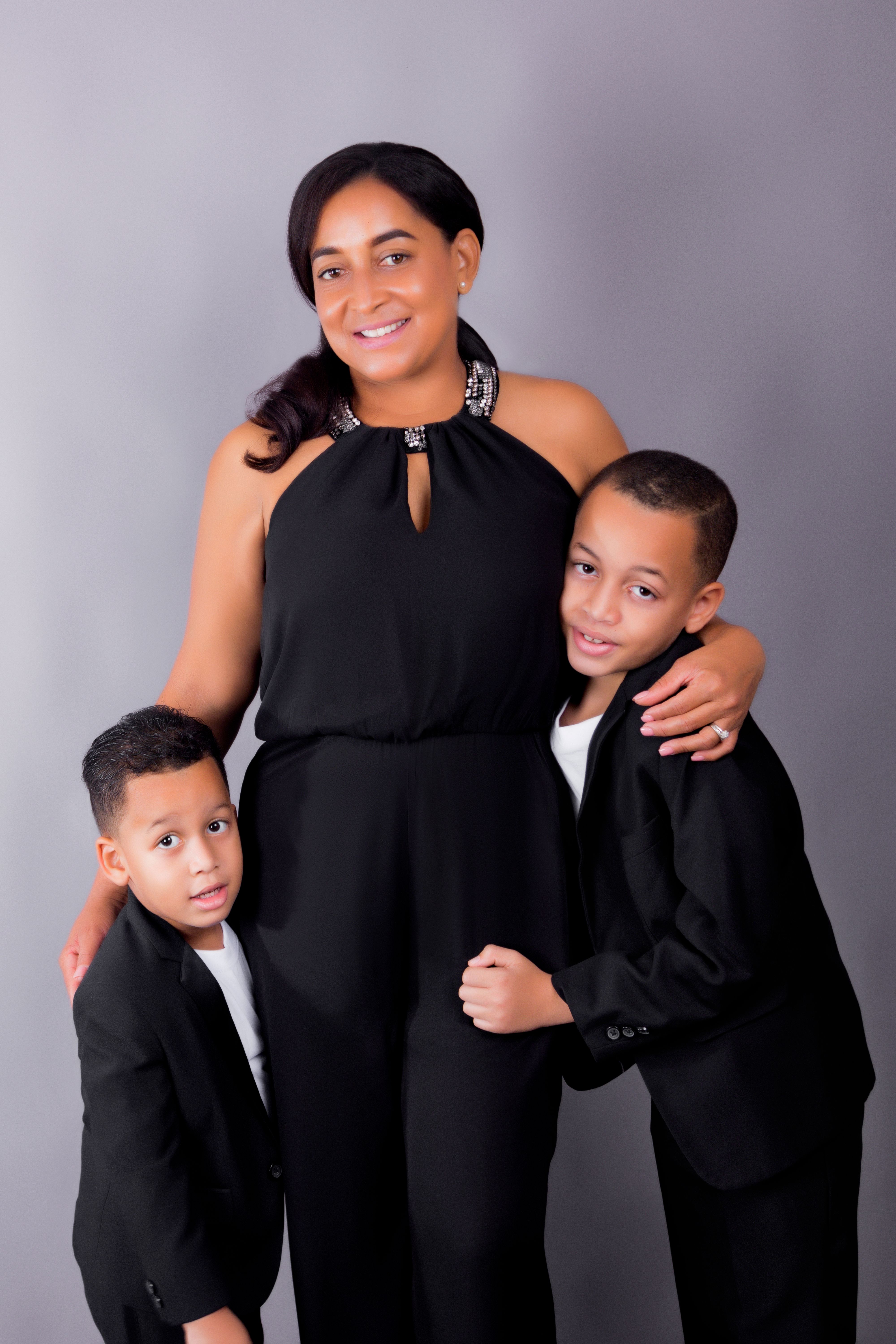 Elegant family portrait with mother and two sons wearing black outfits against a gray studio background.