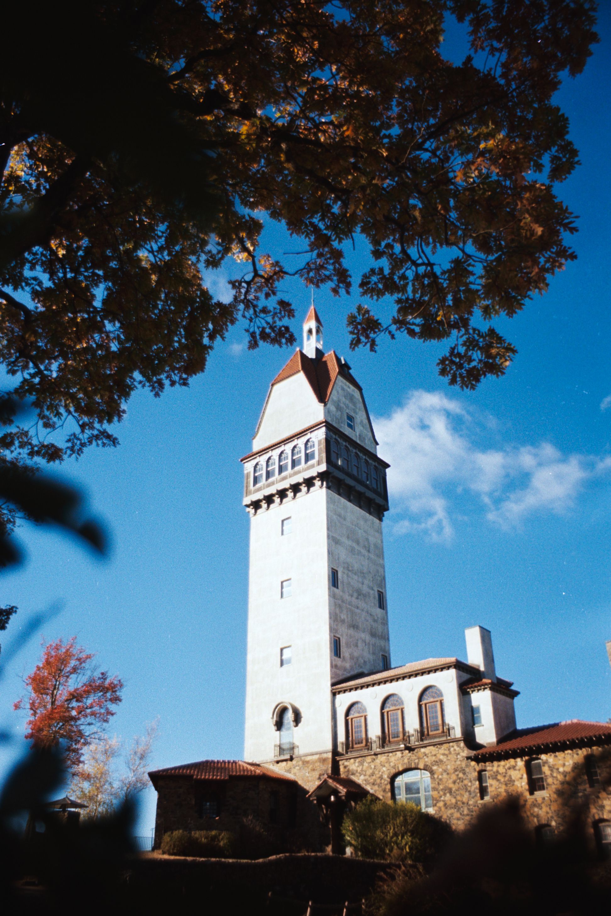 Architectural shot of a stone tower with a red roof surrounded by autumn leaves against a bright blue sky.