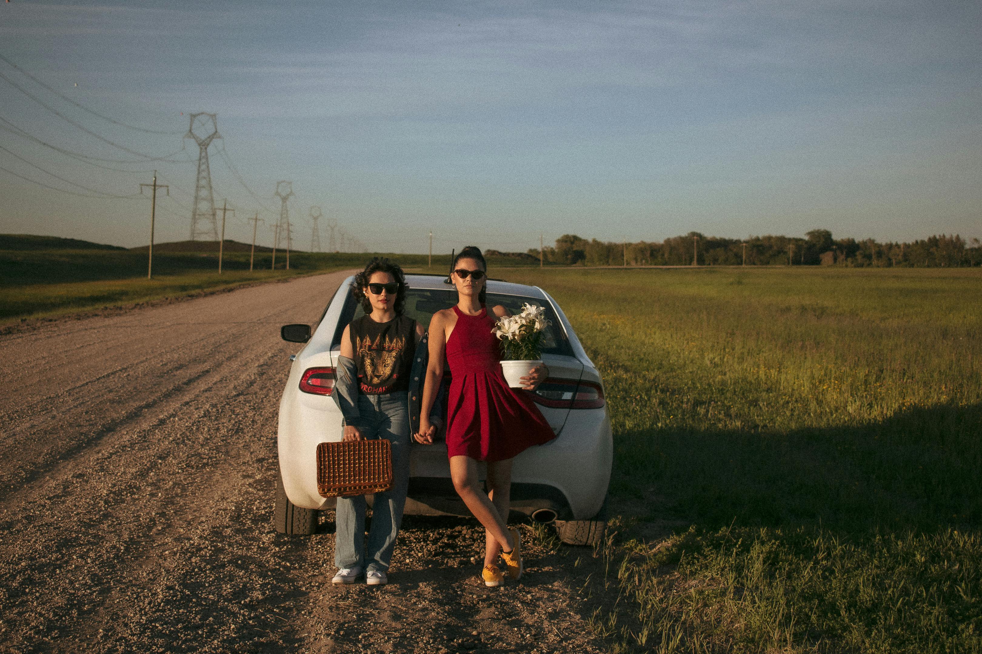 Two women standing by a car on a rural road holding flowers and a basket during a sunny day.