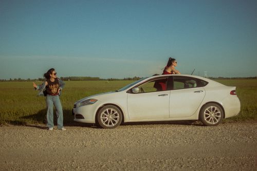 Free Two women enjoying a sunny day on a countryside road with a white car, embracing freedom. Stock Photo