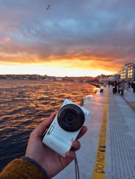 Hand holding a camera with a stunning sunset over the ocean and city skyline.
