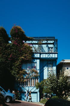 A modern blue house with lush greenery under a clear blue sky, showcasing urban architecture.