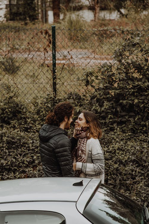 Side view of cheerful woman in outerwear standing near unrecognizable bearded ethnic boyfriend near automobile while looking at each other near trees in daylight