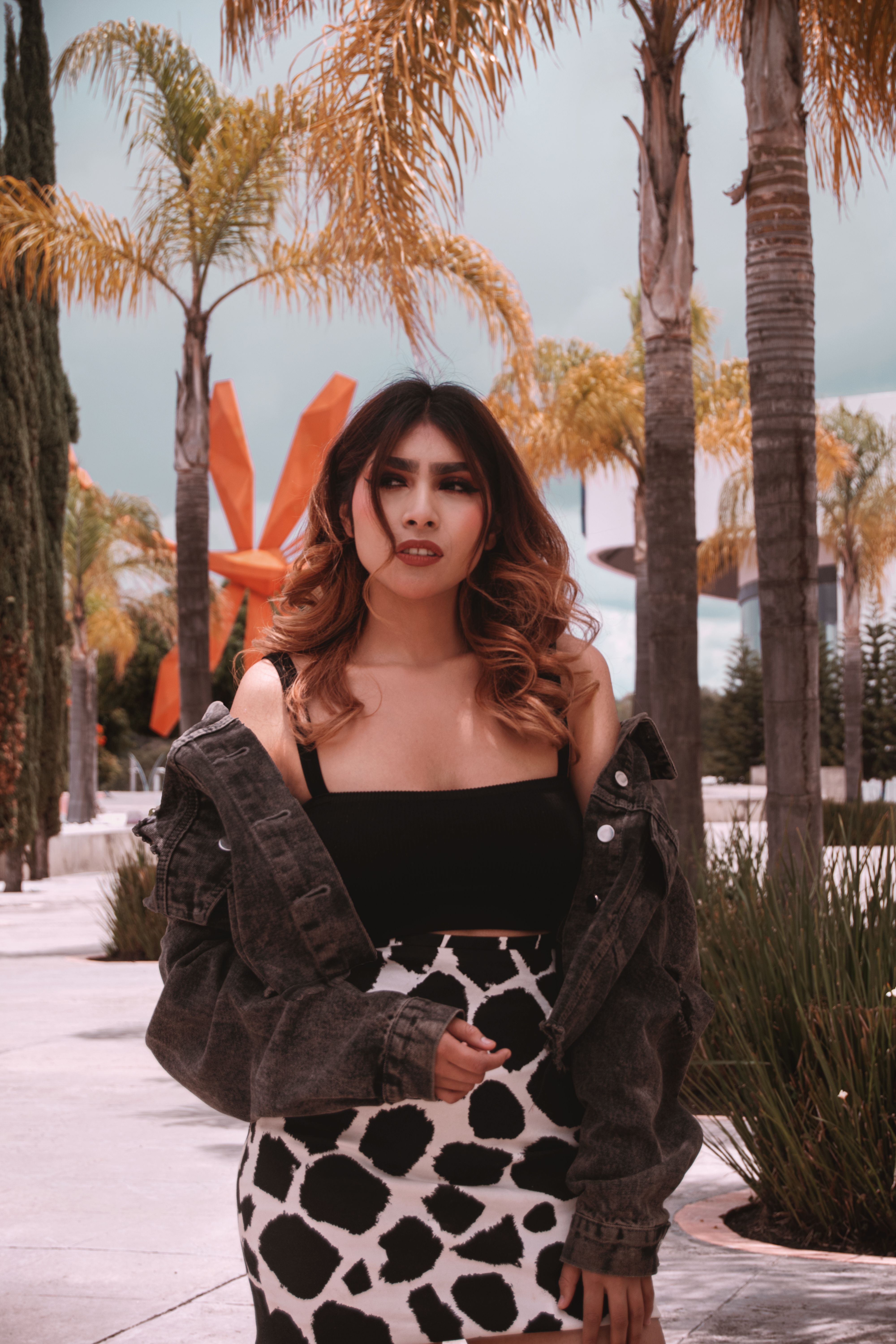 A fashionable woman in an urban park with palm trees and modern art sculpture.