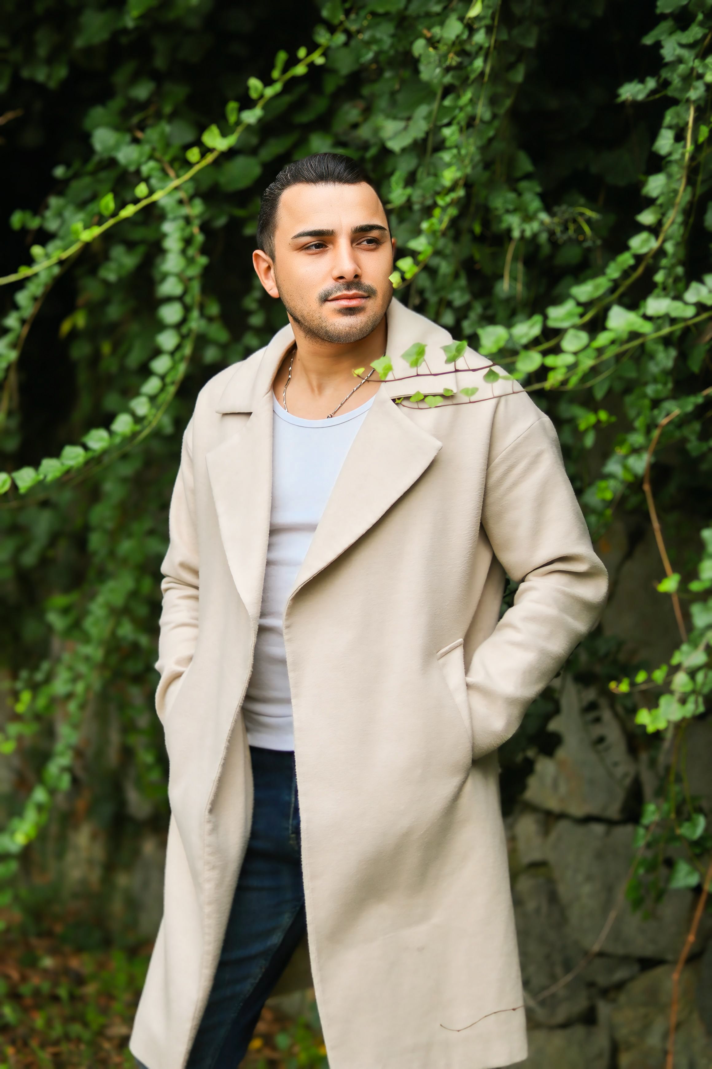 A fashionable man wearing a beige overcoat stands outdoors amid green foliage.