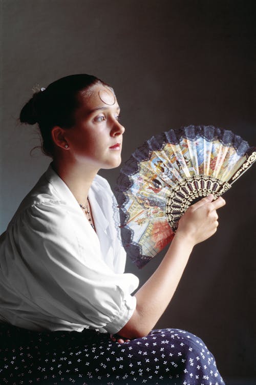 Side view tranquil young female feeling overheated and waving hand fan against face while cooling skin and looking away pensively in dark hot room