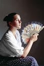 Side view tranquil young female feeling overheated and waving hand fan against face while cooling skin and looking away pensively in dark hot room