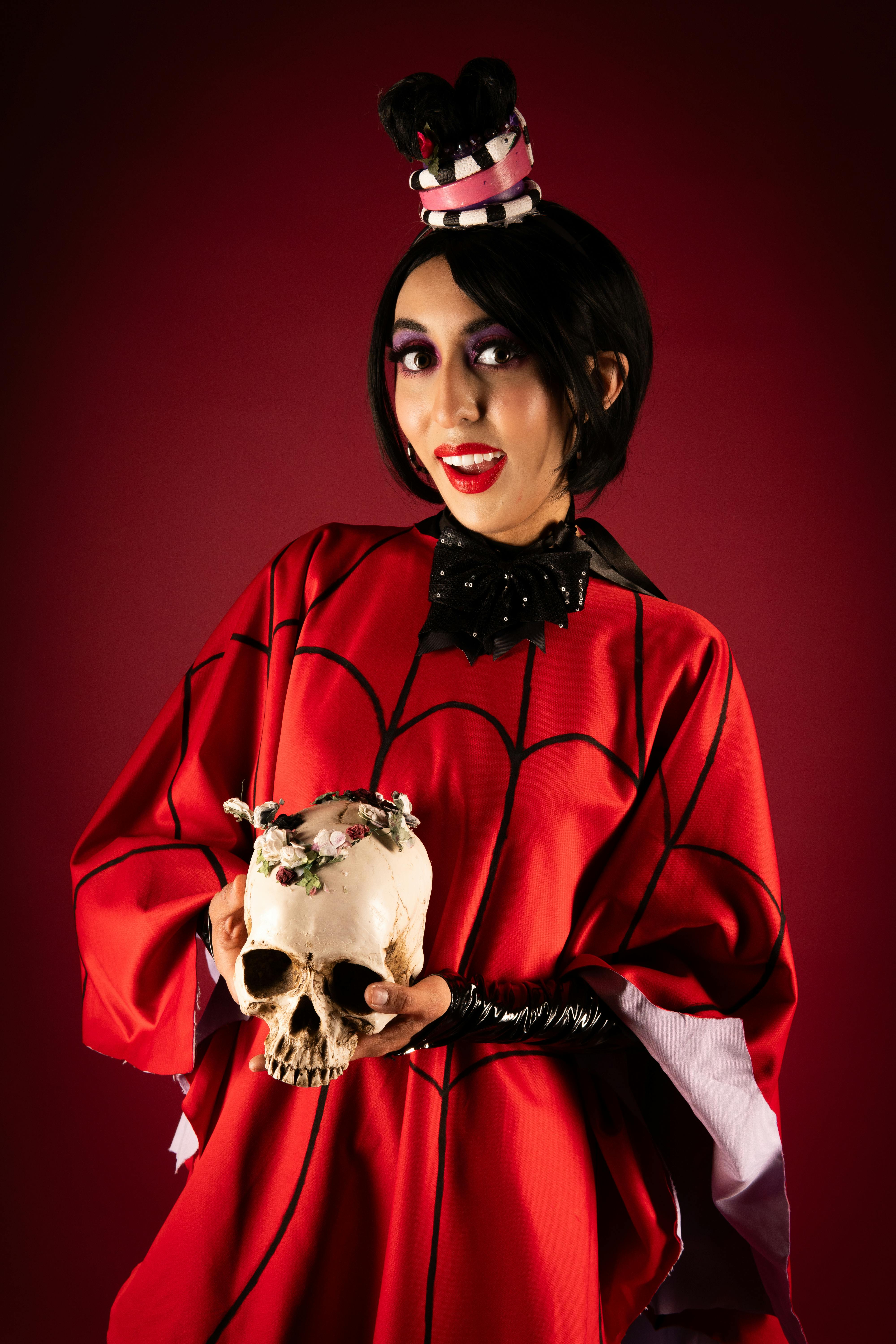 A character in vibrant red cosplay attire holding a decorative skull, against a bold red background.