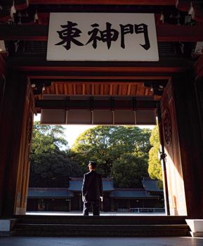 A lone guard stands at the entrance of a Japanese temple, surrounded by lush trees.