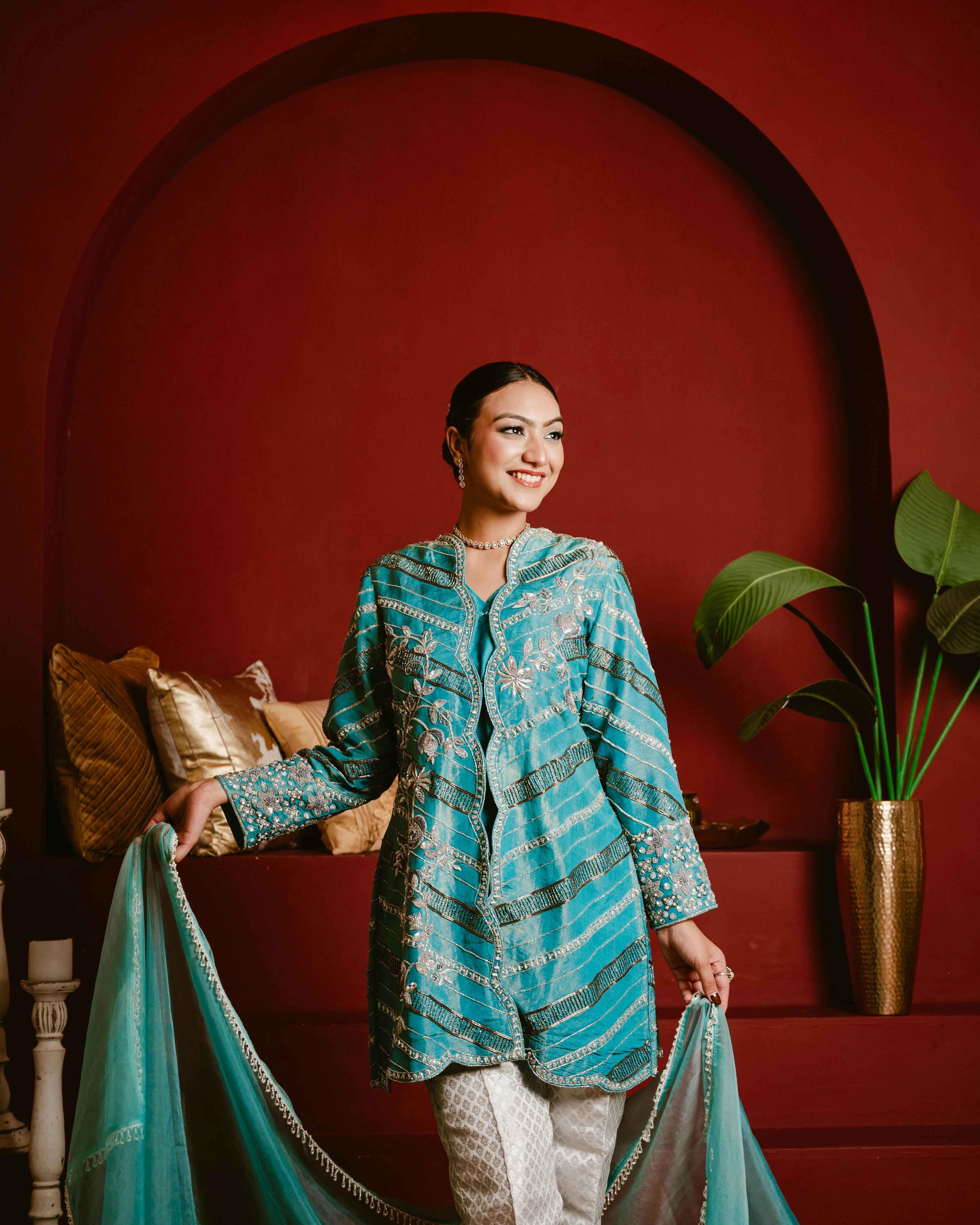 A woman in a stylish blue traditional outfit stands indoors against a rich red background.