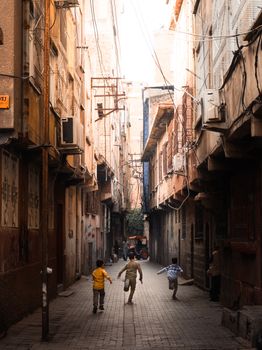 Kids joyfully run through a narrow urban alley, surrounded by rustic buildings.