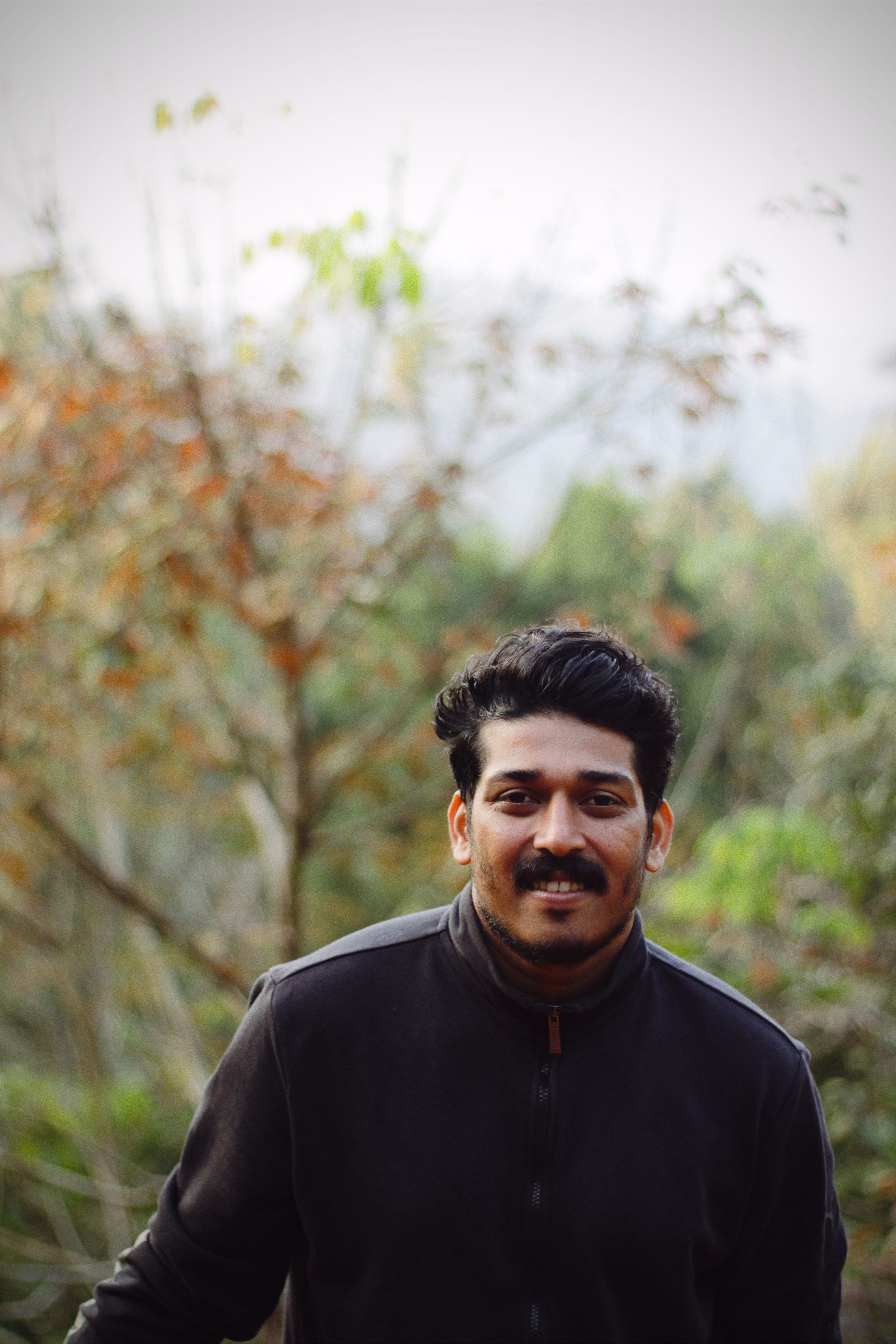 A smiling young South Asian man in a black jacket enjoying the outdoors in fall.