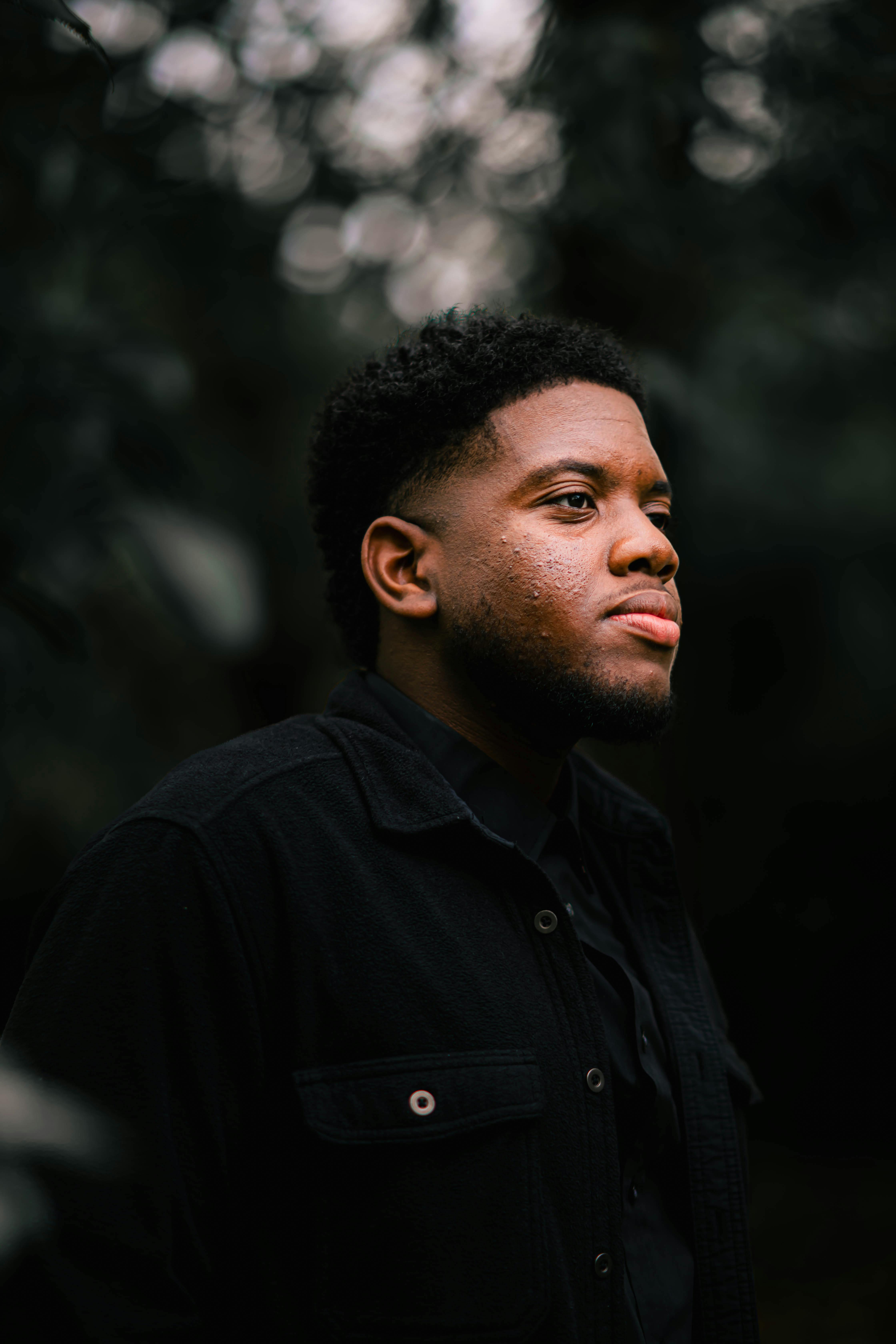 A contemplative young man standing outdoors with a serene expression and natural backdrop.