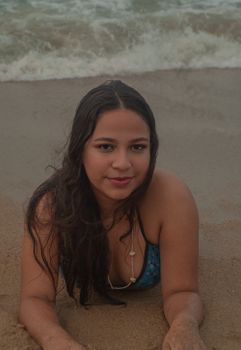 A young woman in a blue swimsuit lying on sandy beach with ocean waves in the background.