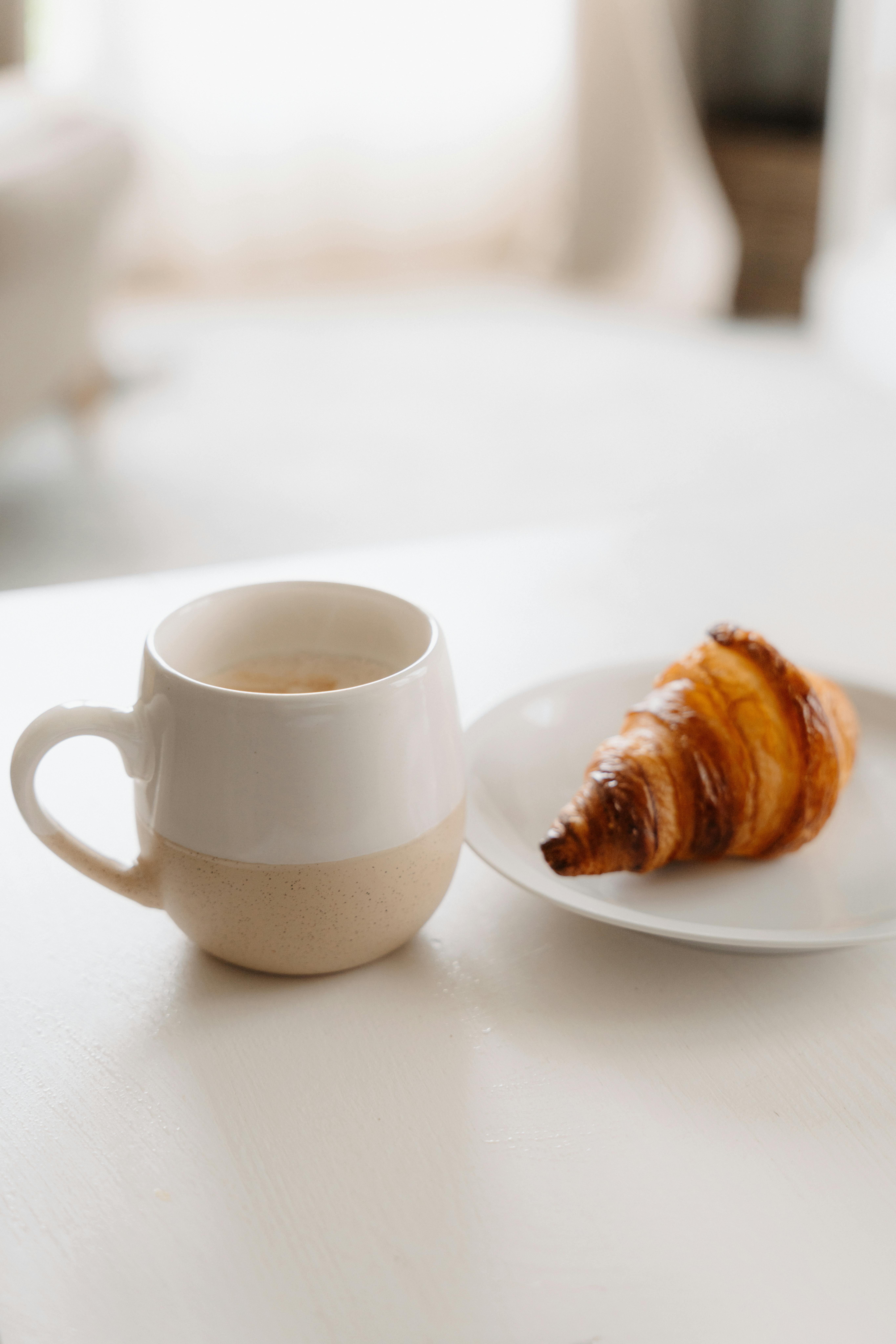 A warm cup of coffee with a fresh croissant on a plate, perfect for a cozy morning snack.