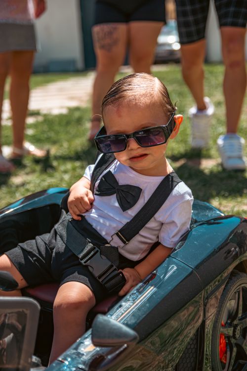 Free Toddler wearing sunglasses and bow tie sitting in a toy car outdoors. Stock Photo