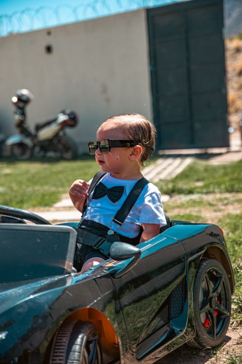 Free Adorable toddler driving a toy car outdoors, wearing sunglasses and a bow tie. Stock Photo