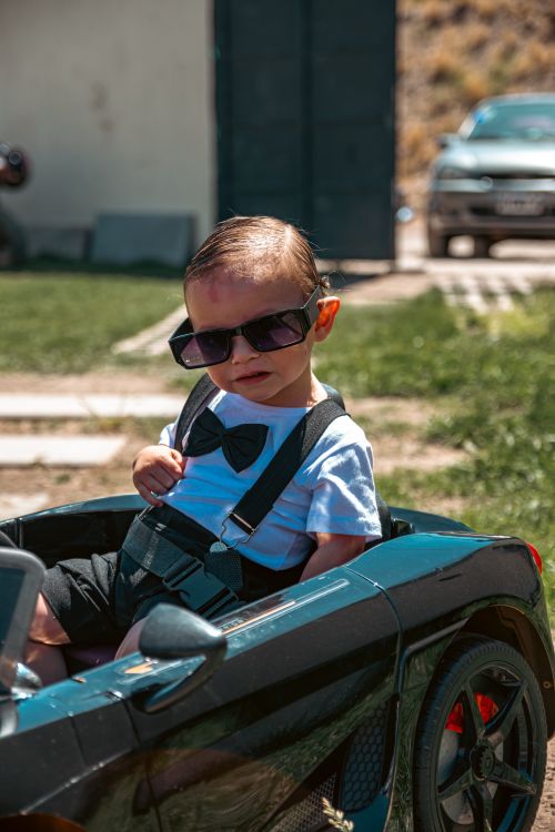 Free Adorable baby dressed in a bow tie and sunglasses sitting in a toy car outdoors. Stock Photo