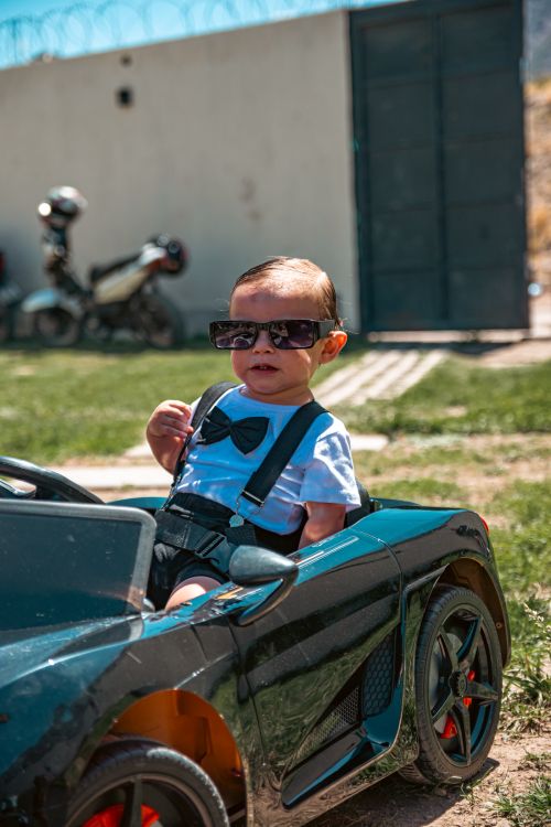 Free A fashionable baby wearing sunglasses rides in a toy sports car outdoors on a sunny day. Stock Photo