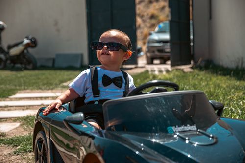 Free A fashionable baby wearing sunglasses drives a toy car in a sunny outdoor setting. Stock Photo