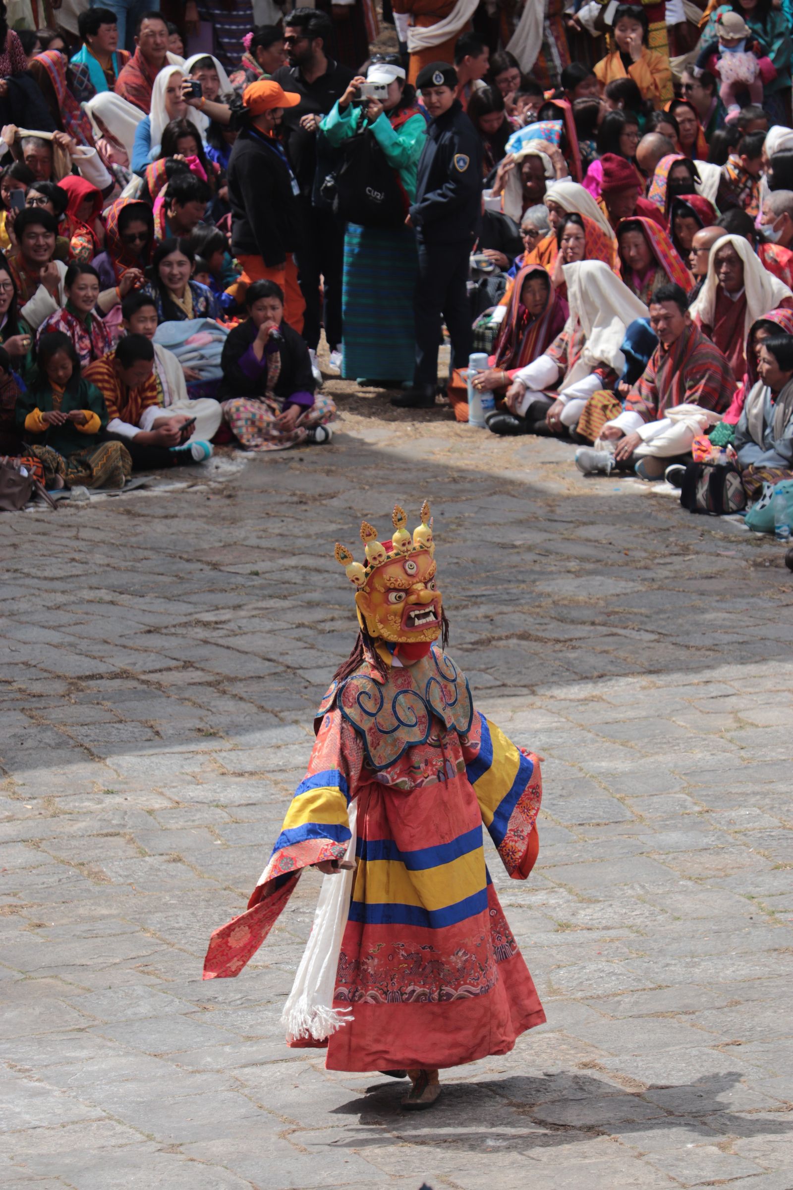 A masked cham dancer in a crowned gold mask and brocade robes turns alone on the paved festival ground, the crowd seated behind