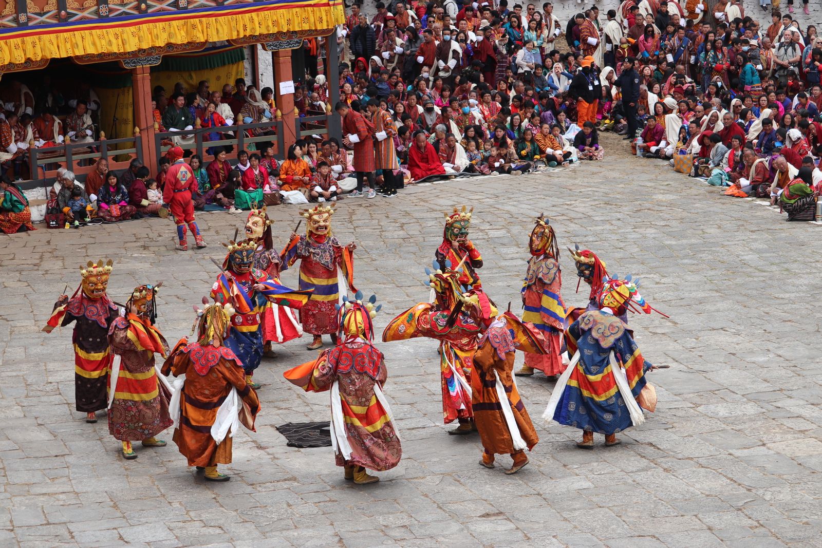 Masked cham dancers in brocade and crowned masks circle a paved festival ground in Bhutan in spring, the crowd seated behind them