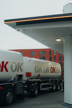 A fuel tanker truck refueling at a contemporary gas station with urban backdrop.