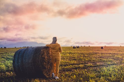 Free A woman stands by a hay bale in a vast field during a tranquil sunset. Stock Photo