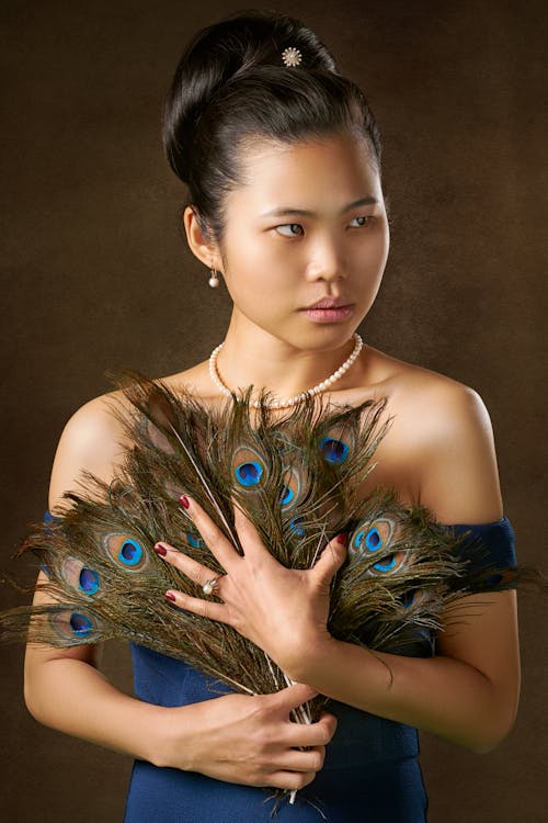 Photo of Woman Standing In Front of Brown Background While Holding Peacock Feathers