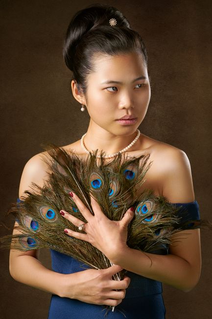 Sophisticated portrait of a woman in blue attire holding peacock feathers, posed against a brown bac