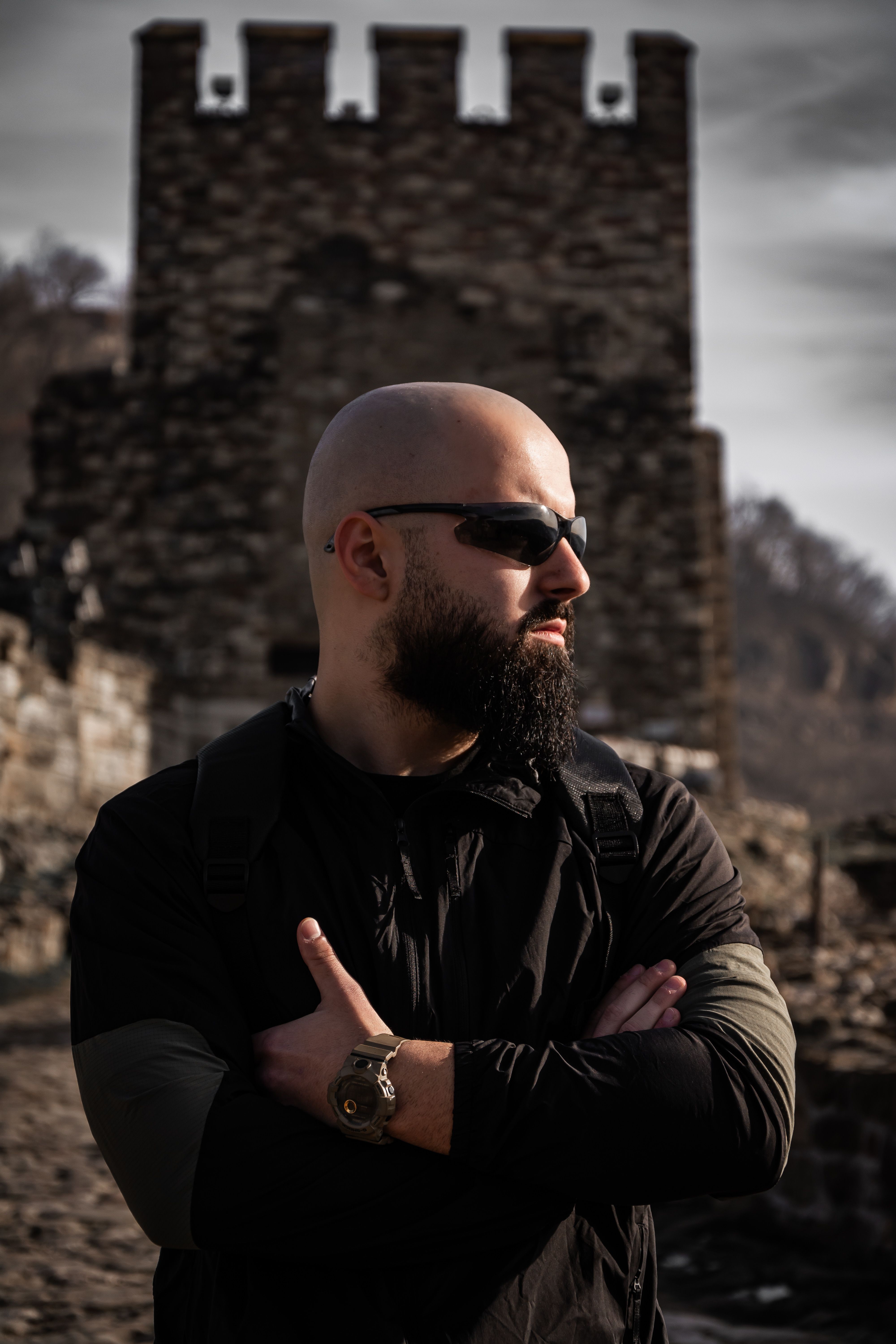 Bearded man in sunglasses posing confidently outdoors near a historic stone tower.
