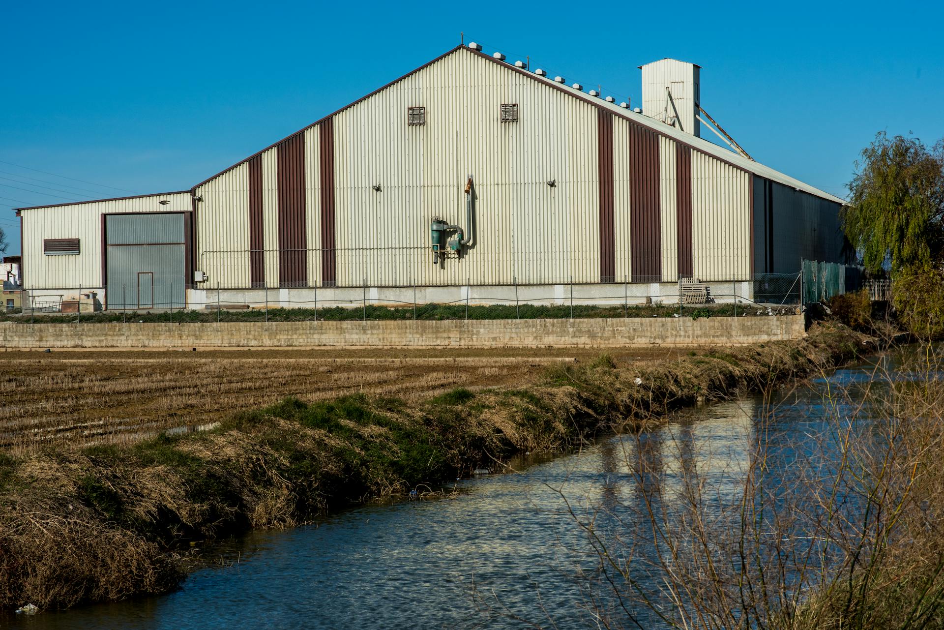 A large industrial warehouse stands next to a serene creek during the day.