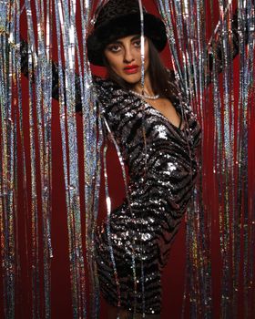 A stylish woman in a sequin dress and hat poses amidst shimmering streamers against a red backdrop.