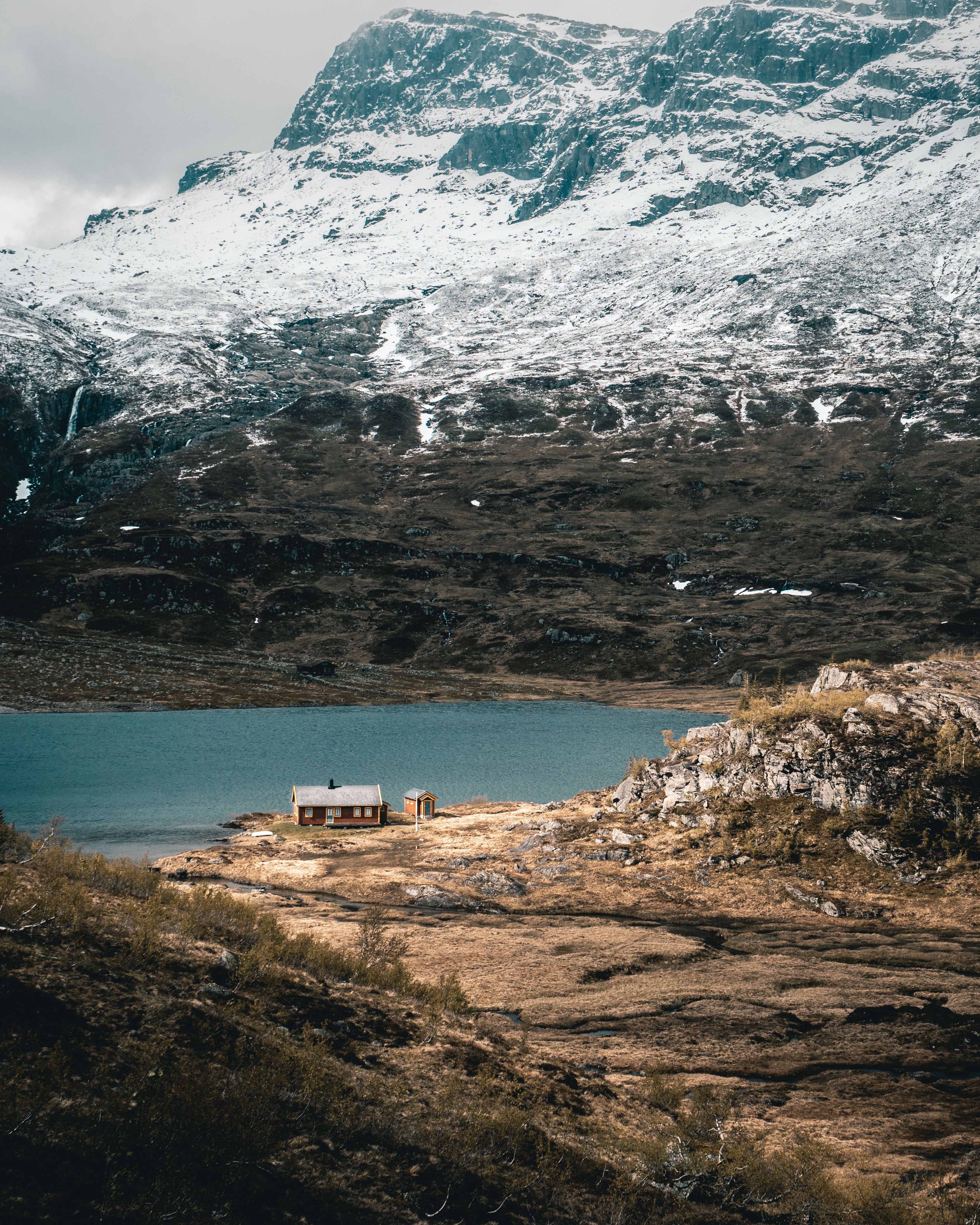 A lone cabin rests beside a serene mountain lake surrounded by snowy peaks.