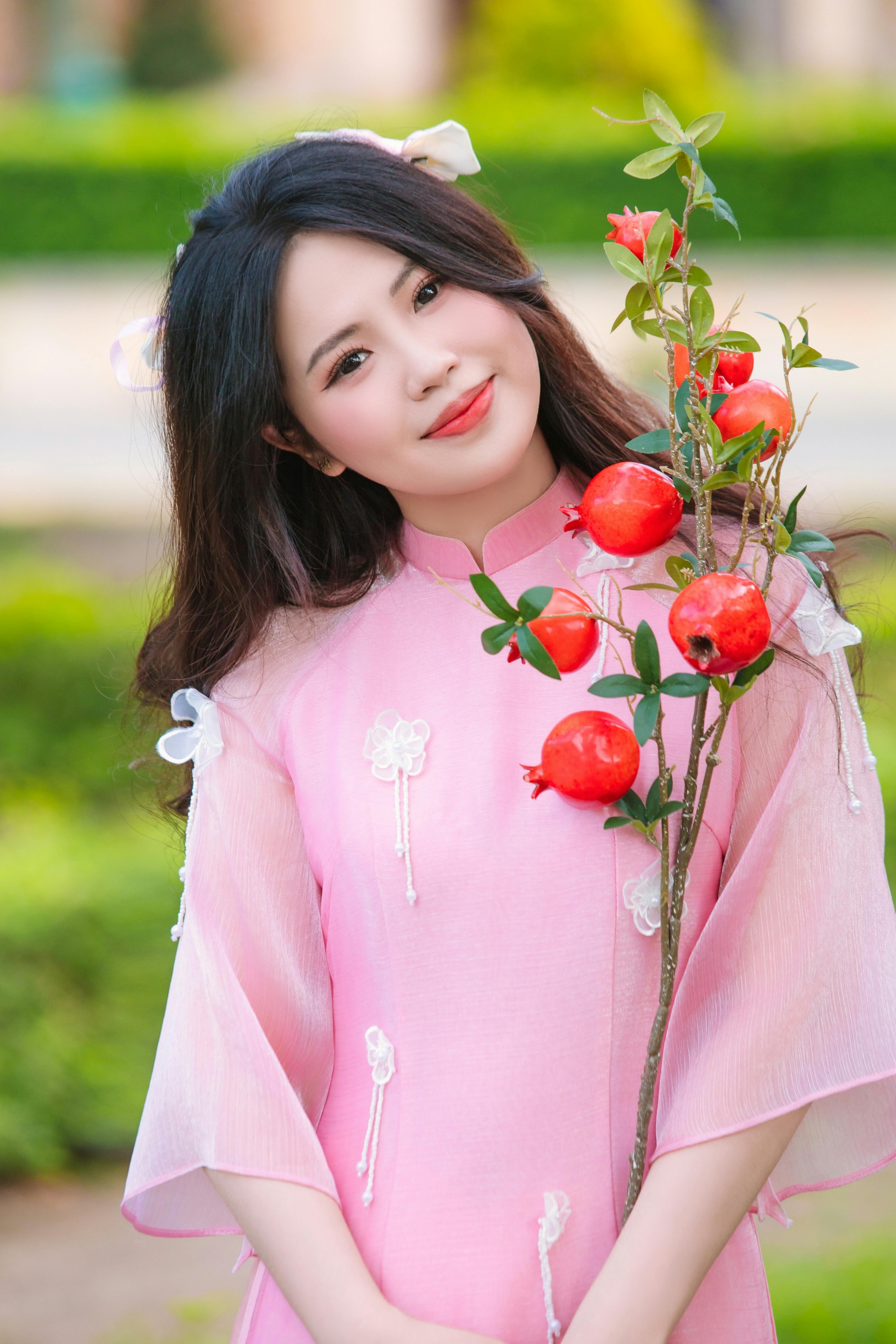 Smiling young woman holding a pomegranate branch, wearing a traditional pink dress outdoors.