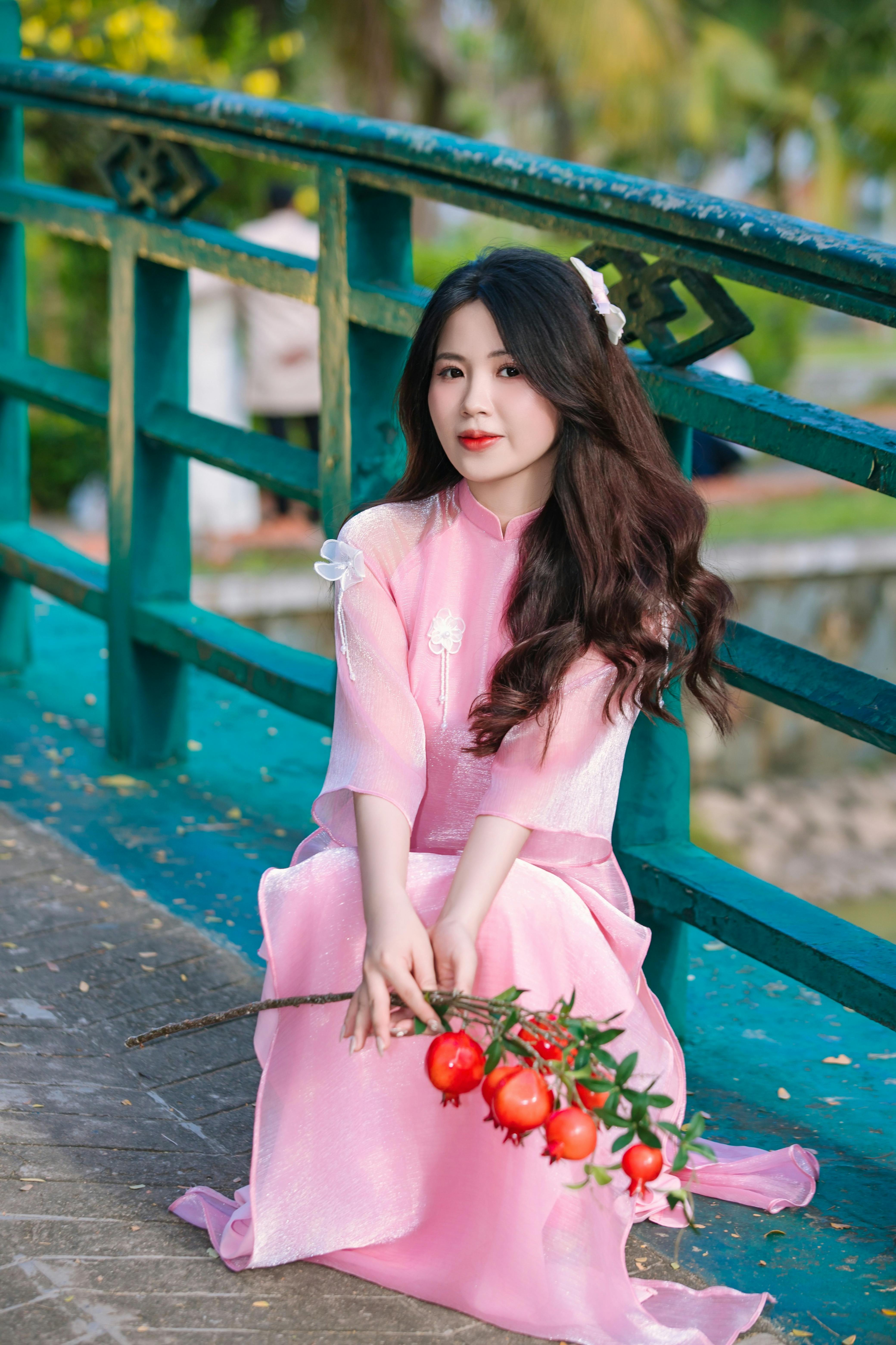 Graceful woman wearing pink dress with long hair sitting on bridge holding fruit branch.