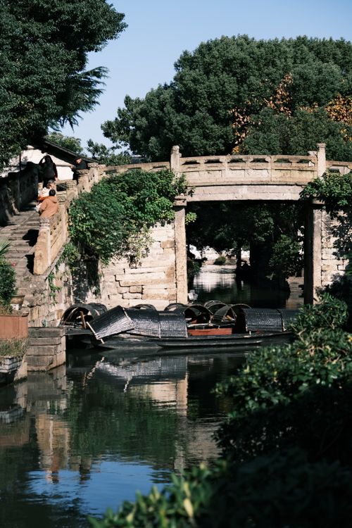 Free Scenic view of a traditional stone bridge over a peaceful waterway with boats and lush greenery. Stock Photo