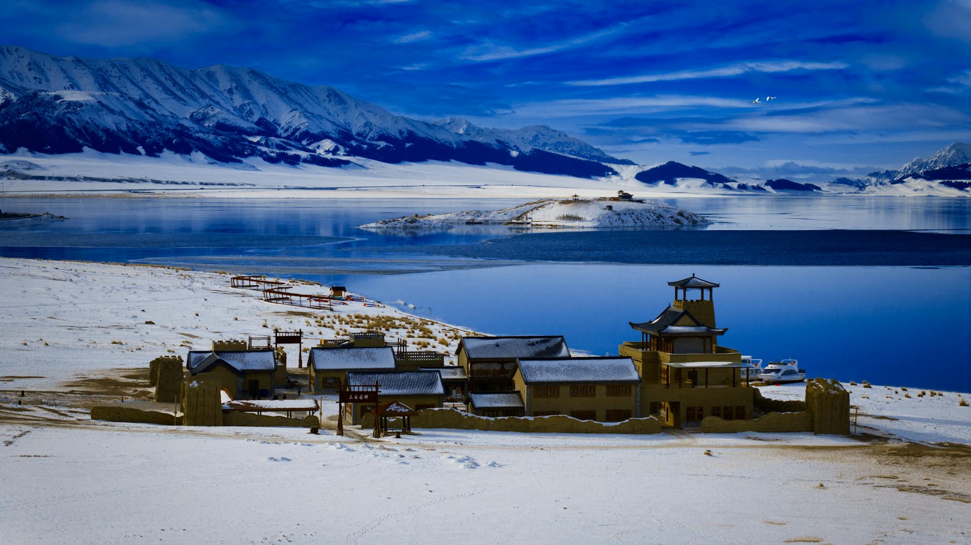 Snow-covered village by tranquil lake with mountains in the background, a serene winter scene.