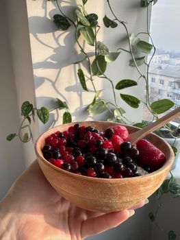 Healthy mixed berries in a wooden bowl with natural light, wooden spoon, green leaves.