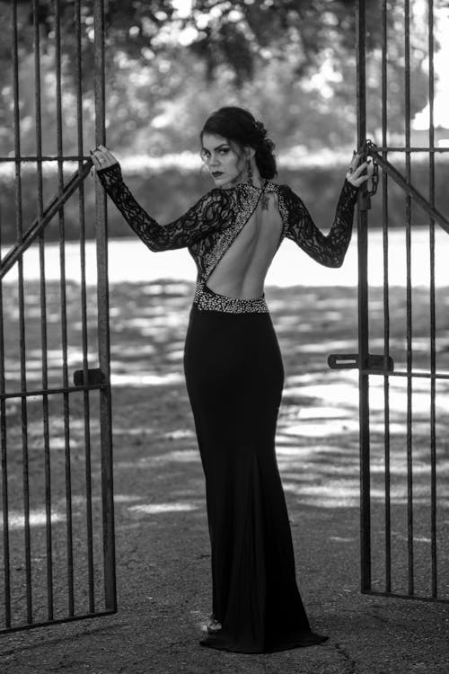 Фотография женщины в элегантном платье позирует у металлических ворот, оглядываясь назад, в оттенках серого