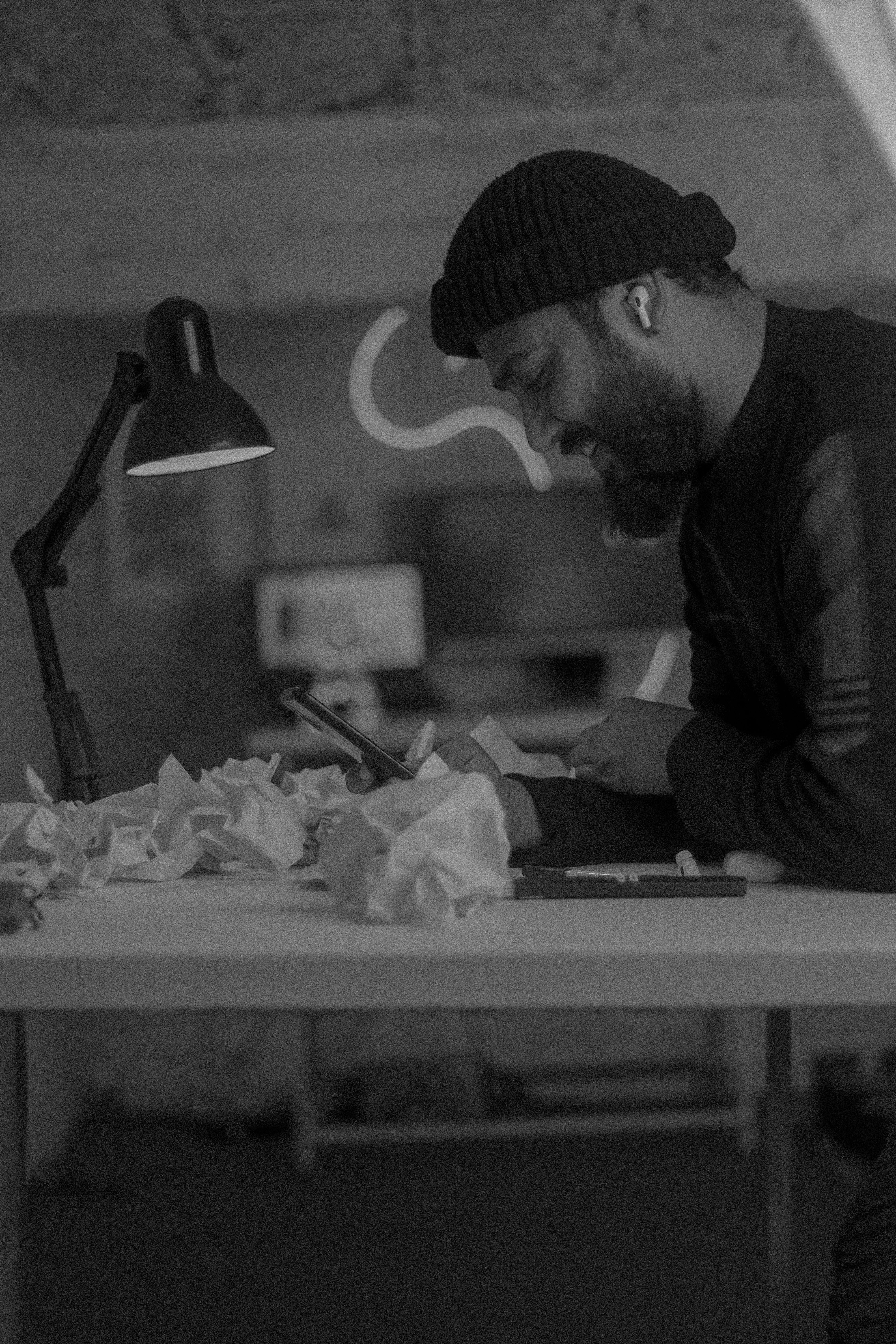 Bearded man in beanie works intently at cluttered desk under lamp light, surrounded by crumpled papers.