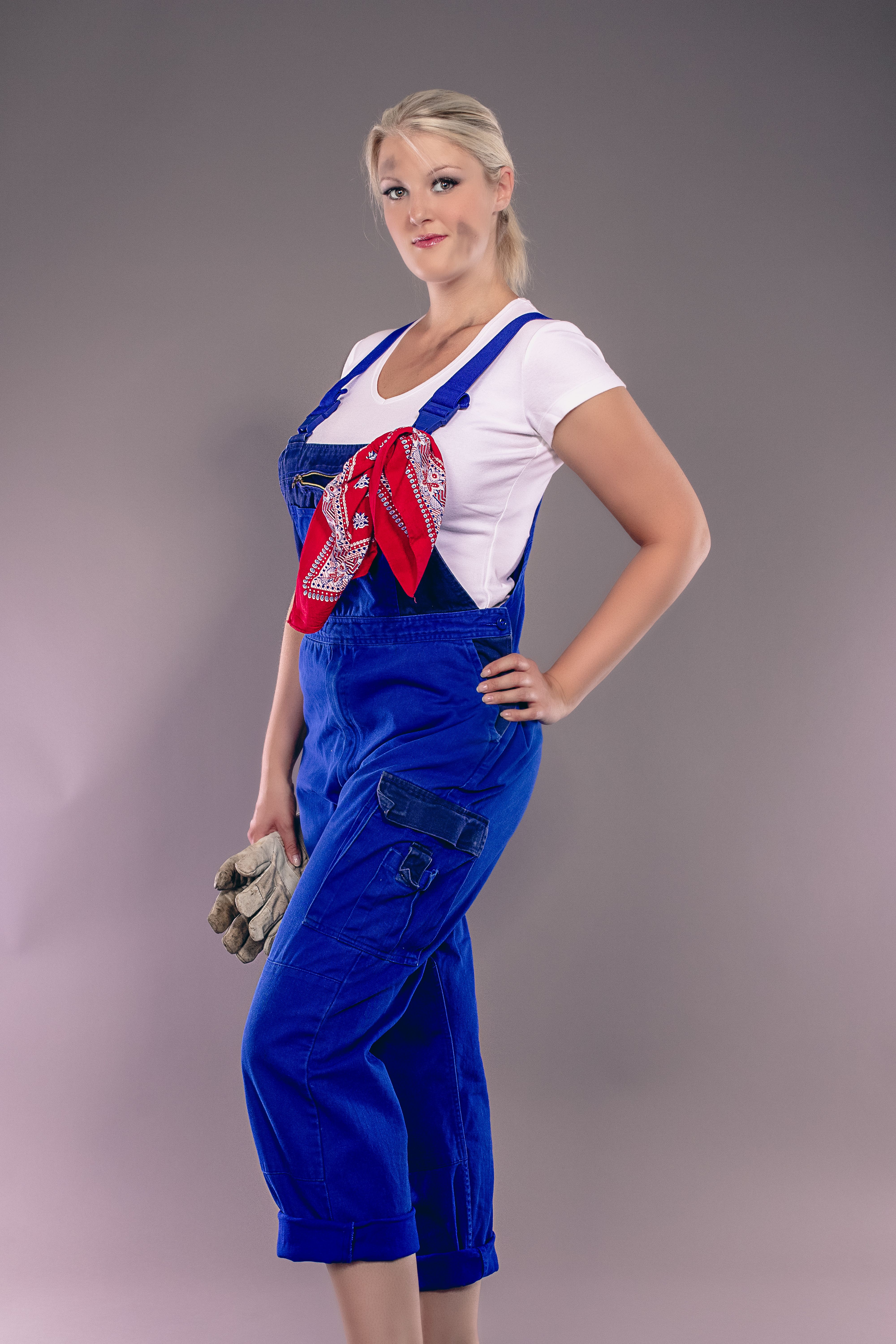 Confident woman in work overalls with gloves and red bandana, studio background.
