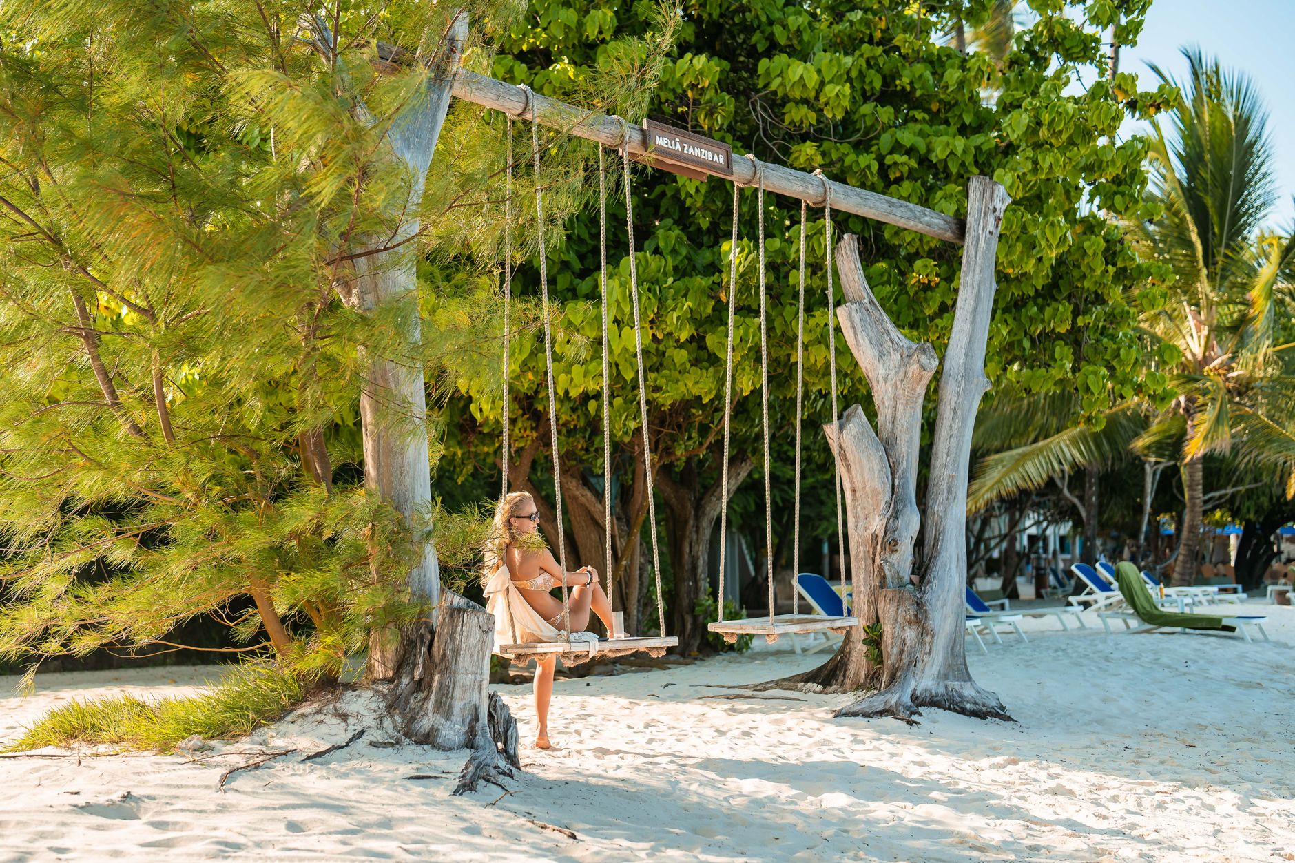 Woman enjoying a sunny day on a beach swing beneath palm trees in Zanzibar.