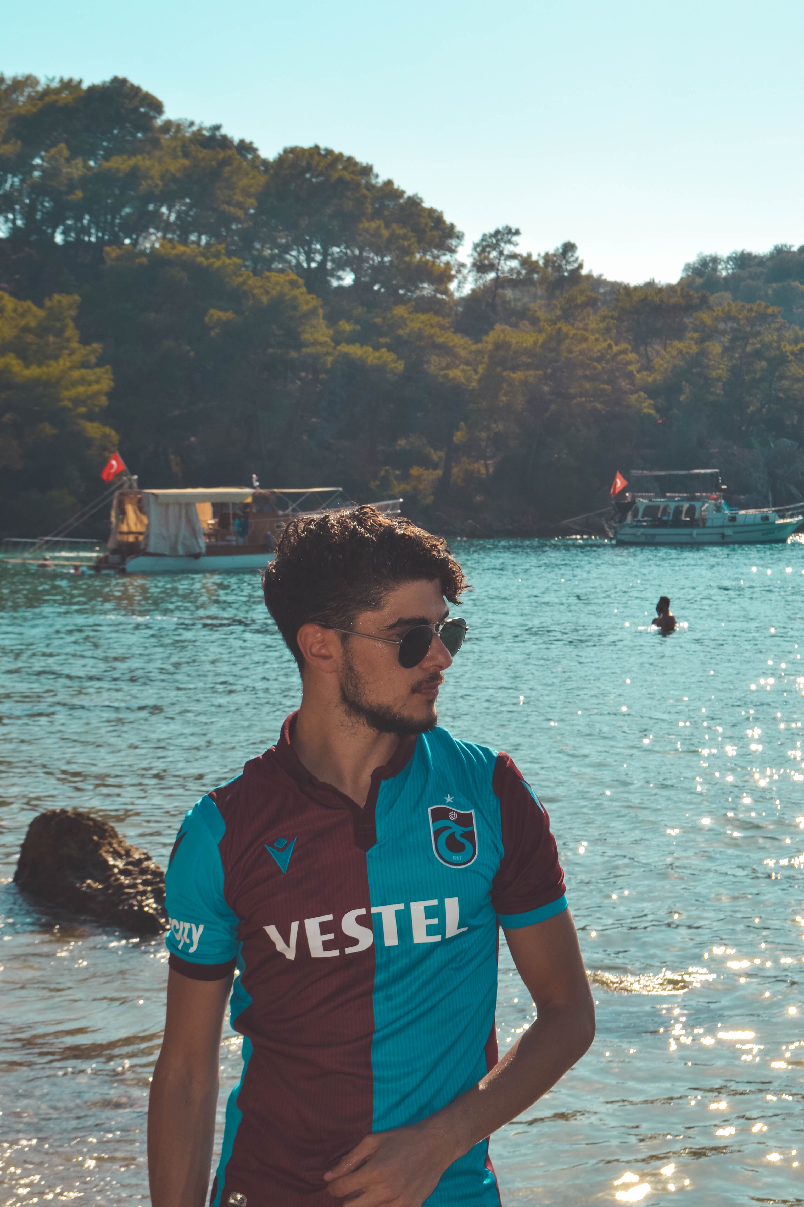 Man in a football jersey by a waterfront, surrounded by boats and trees.