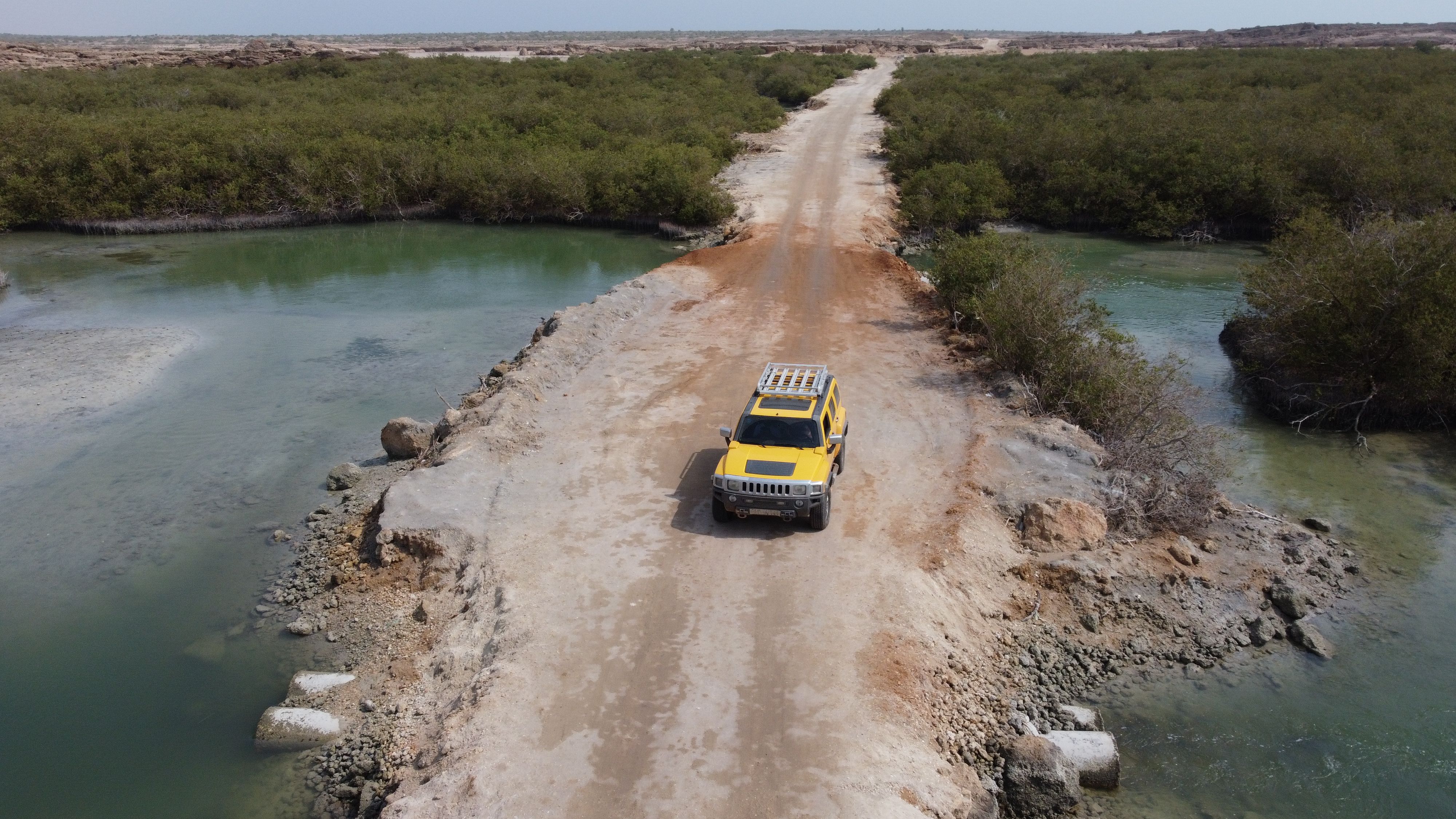 Aerial view of a yellow SUV crossing a remote desert road with surrounding greenery and water.
